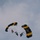 USAPT Golden Knights Parachutist Jump into Kentucky Derby Two military skydivers are descending under black and gold parachutes marked with 'U.S. ARMY' branding, consistent with the U.S. Army Golden Knights parachute team. One jumper is carrying an American flag during the descent. The image is taken from a low angle against an overcast sky.