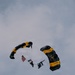 Two military skydivers are descending under black and gold parachutes marked with 'U.S. ARMY' branding, consistent with the U.S. Army Golden Knights parachute team. One jumper is carrying an American flag during the descent. The image is taken from a low angle against an overcast sky.