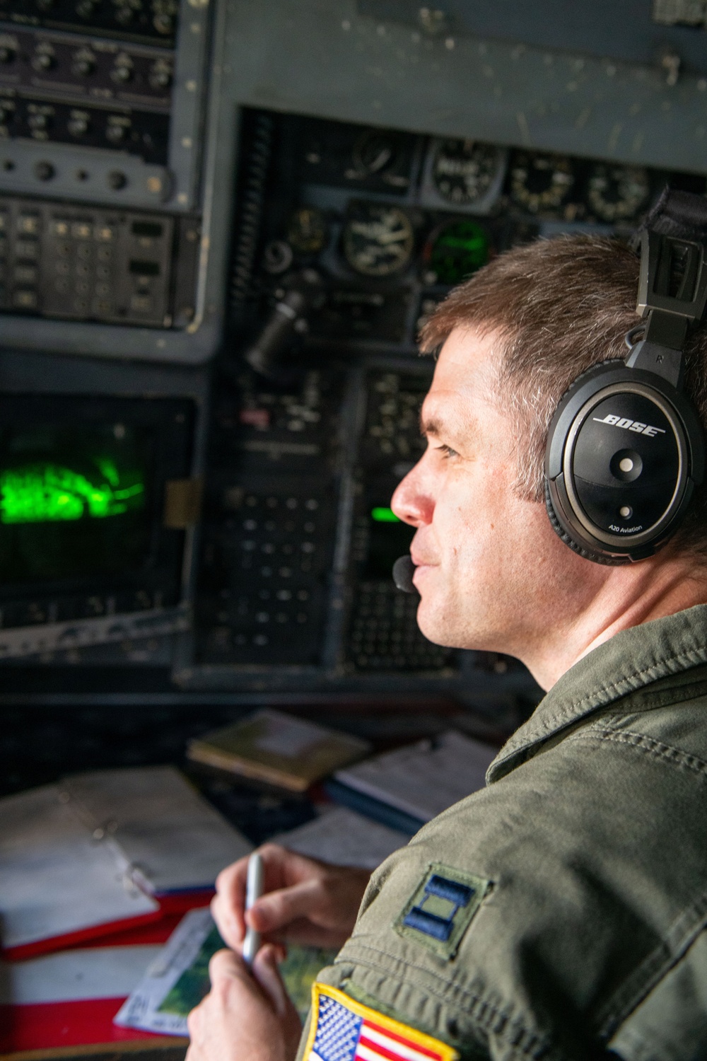A U.S. military aviator is seated in an aircraft cockpit, wearing a Bose A20 Aviation headset and an olive green flight suit with a U.S. flag patch and captain rank insignia. He is holding a pen and appears to be monitoring instruments or looking out the cockpit window. The background shows a complex array of analog and digital cockpit instruments, including a green-lit display screen.