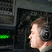 A U.S. military aviator is seated in an aircraft cockpit, wearing a Bose A20 Aviation headset and an olive green flight suit with a U.S. flag patch and captain rank insignia. He is holding a pen and appears to be monitoring instruments or looking out the cockpit window. The background shows a complex array of analog and digital cockpit instruments, including a green-lit display screen.