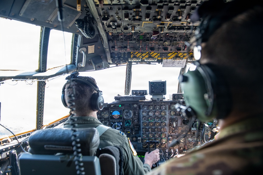 Two military aircrew members are seated in the cockpit of a large military transport aircraft, likely a C-130 Hercules, during flight. Both crew members are wearing flight suits and aviation headsets, facing an extensive analog and digital instrument panel. The aircraft appears to be airborne based on the view through the windscreen.