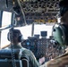 Two military aircrew members are seated in the cockpit of a large military transport aircraft, likely a C-130 Hercules, during flight. Both crew members are wearing flight suits and aviation headsets, facing an extensive analog and digital instrument panel. The aircraft appears to be airborne based on the view through the windscreen.