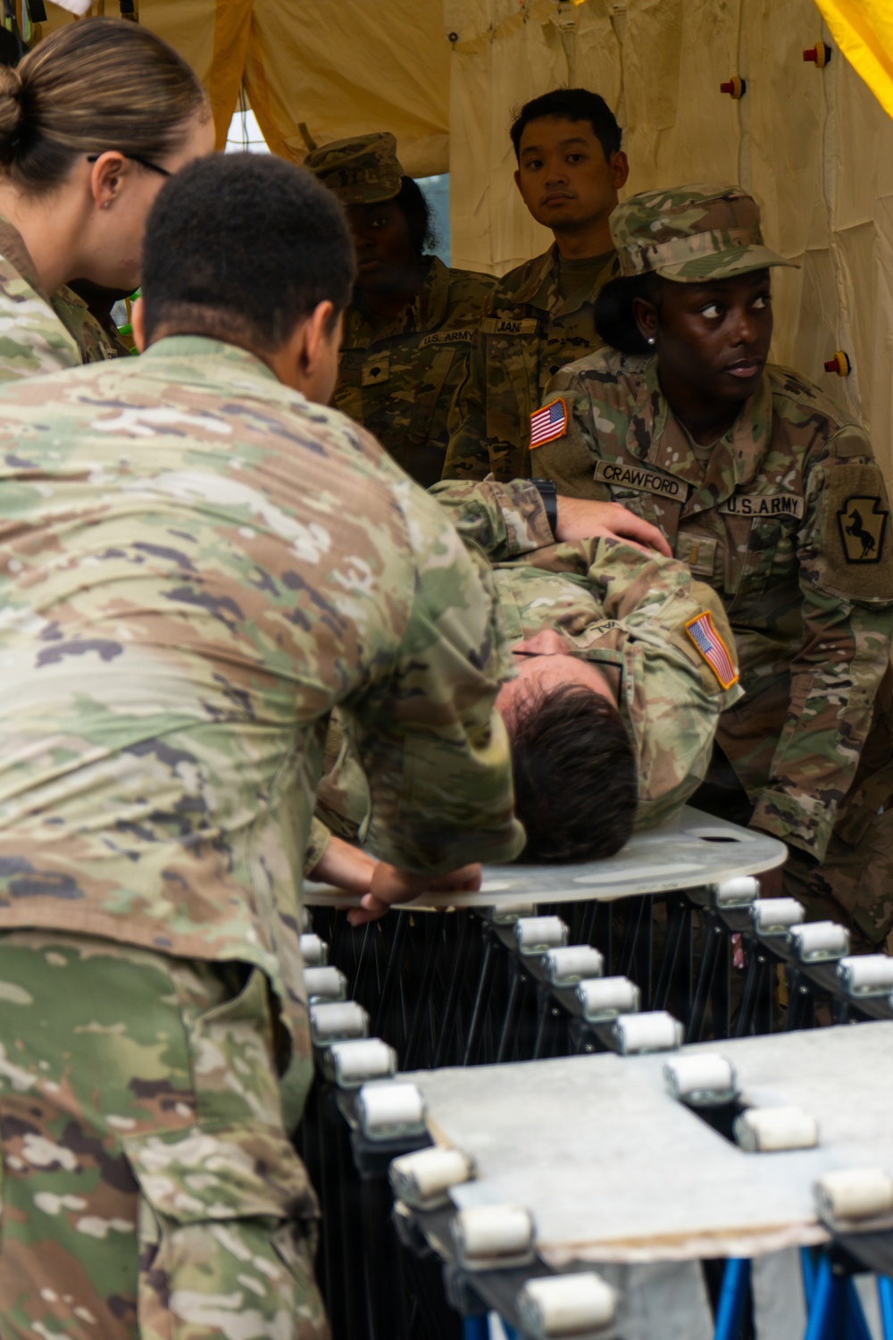 U.S. Army soldiers are conducting a medical training exercise inside a military field tent. A simulated casualty is being transported on a litter/stretcher by multiple soldiers, while others observe the procedure. Name tapes visible include 'CRAWFORD' and 'JAN', and soldiers wear OCP (Operational Camouflage Pattern) uniforms with U.S. Army patches.