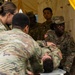 U.S. Army soldiers are conducting a medical training exercise inside a military field tent. A simulated casualty is being transported on a litter/stretcher by multiple soldiers, while others observe the procedure. Name tapes visible include 'CRAWFORD' and 'JAN', and soldiers wear OCP (Operational Camouflage Pattern) uniforms with U.S. Army patches.