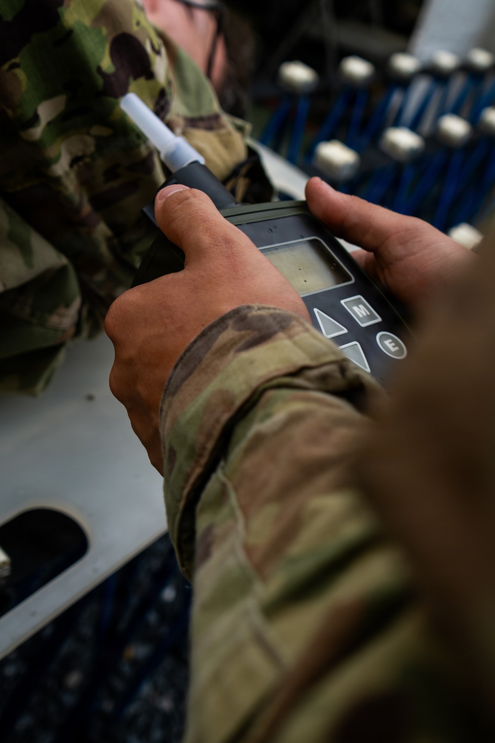 A military service member in OCP (Operational Camouflage Pattern) uniform is holding a handheld electronic detection device with a probe/wand attachment and a small LCD display with navigation buttons. The device appears to be a chemical, biological, radiological, or nuclear (CBRN) detection instrument or similar field measurement tool. The background shows what appears to be the interior of a military aircraft or vehicle with blue-handled equipment racks.