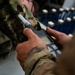 A military service member in OCP (Operational Camouflage Pattern) uniform is holding a handheld electronic detection device with a probe/wand attachment and a small LCD display with navigation buttons. The device appears to be a chemical, biological, radiological, or nuclear (CBRN) detection instrument or similar field measurement tool. The background shows what appears to be the interior of a military aircraft or vehicle with blue-handled equipment racks.