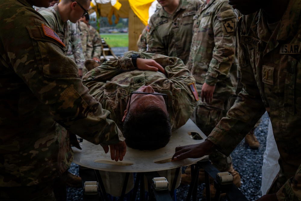 Multiple U.S. Army soldiers in OCP (Operational Camouflage Pattern) uniforms are attending to a fellow soldier lying on a stretcher or litter board. The scene appears to be a medical training exercise or casualty evacuation drill conducted under a yellow tarp or tent structure. Several personnel are positioned around the patient, suggesting coordinated litter-carry or triage procedures.