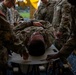 Multiple U.S. Army soldiers in OCP (Operational Camouflage Pattern) uniforms are attending to a fellow soldier lying on a stretcher or litter board. The scene appears to be a medical training exercise or casualty evacuation drill conducted under a yellow tarp or tent structure. Several personnel are positioned around the patient, suggesting coordinated litter-carry or triage procedures.