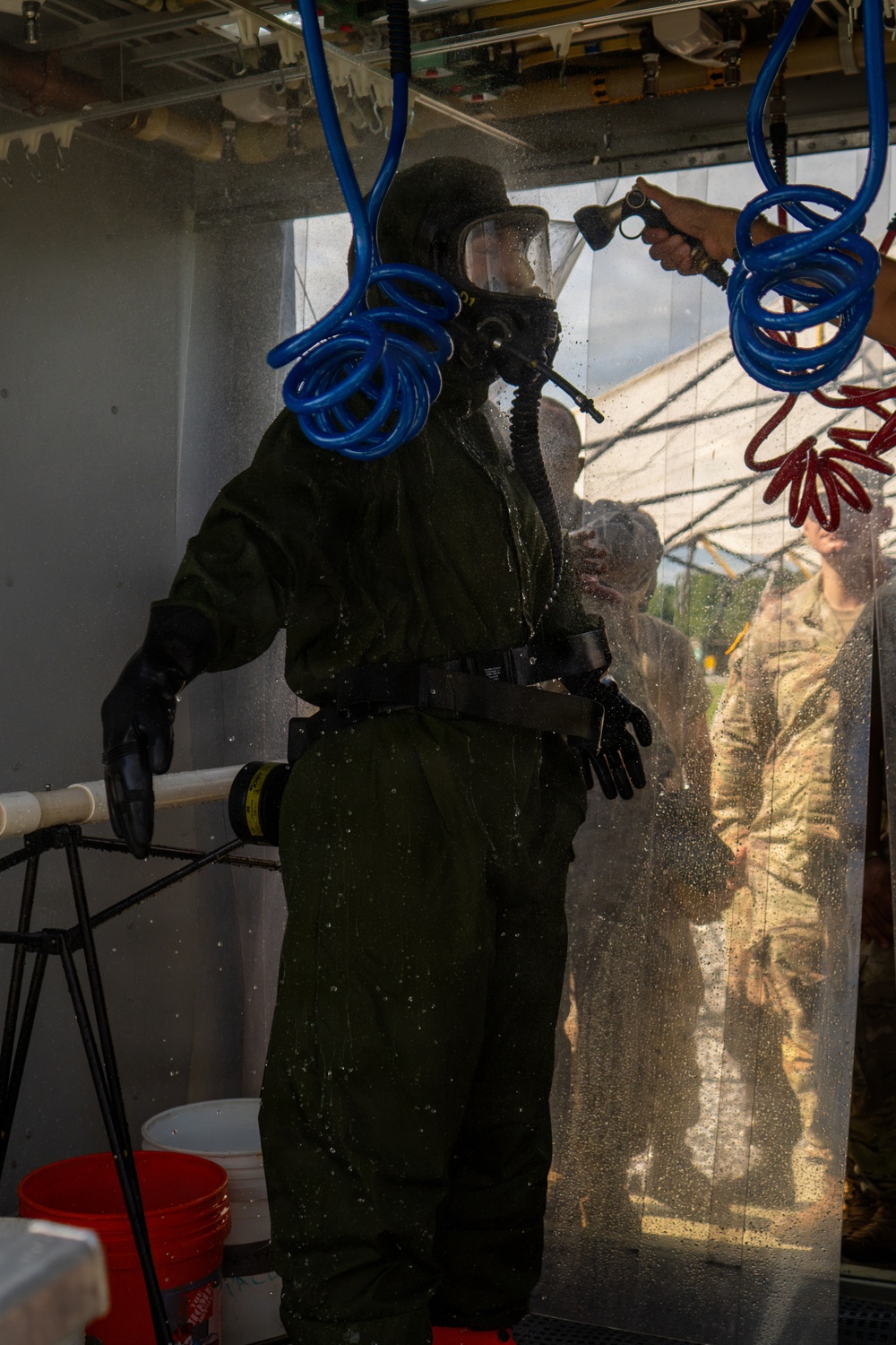 A person wearing a full CBRN (Chemical, Biological, Radiological, and Nuclear) protective suit with an integrated respirator/gas mask is undergoing a decontamination shower procedure. The individual is being sprayed with water by an attendant using a hose nozzle inside what appears to be a decontamination station or tent. Military personnel in camouflage uniforms are visible through a clear plastic curtain barrier in the background.