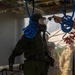 A person wearing a full CBRN (Chemical, Biological, Radiological, and Nuclear) protective suit with an integrated respirator/gas mask is undergoing a decontamination shower procedure. The individual is being sprayed with water by an attendant using a hose nozzle inside what appears to be a decontamination station or tent. Military personnel in camouflage uniforms are visible through a clear plastic curtain barrier in the background.