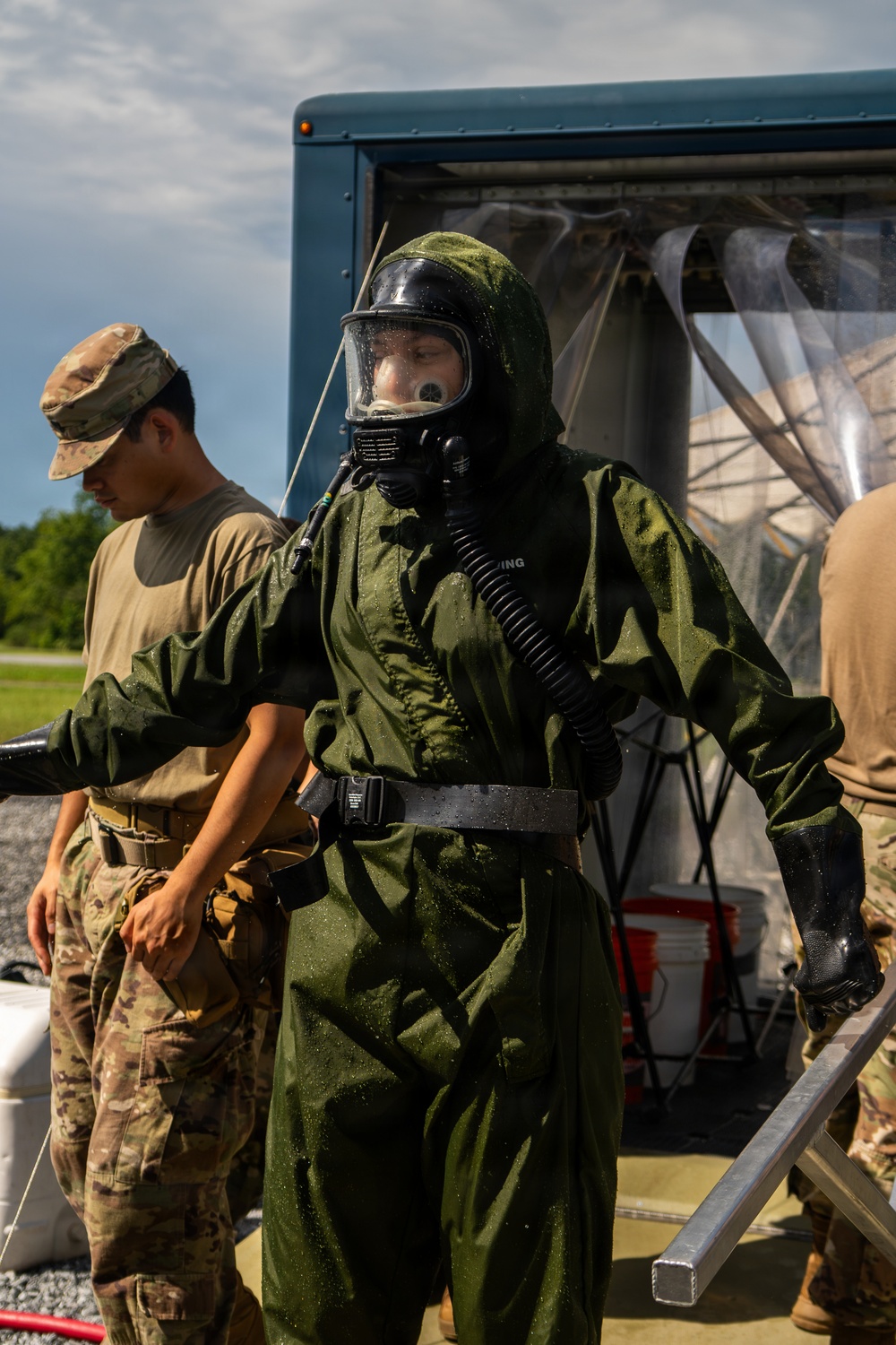 A military personnel is wearing a full CBRN (Chemical, Biological, Radiological, and Nuclear) protective suit with an integrated respirator/gas mask, appearing wet from a decontamination procedure. At least two other soldiers in standard multicam/OCP uniforms are present nearby. The setting appears to be an outdoor military training or exercise area with a trailer and decontamination station visible in the background.