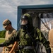 A military personnel is wearing a full CBRN (Chemical, Biological, Radiological, and Nuclear) protective suit with an integrated respirator/gas mask, appearing wet from a decontamination procedure. At least two other soldiers in standard multicam/OCP uniforms are present nearby. The setting appears to be an outdoor military training or exercise area with a trailer and decontamination station visible in the background.