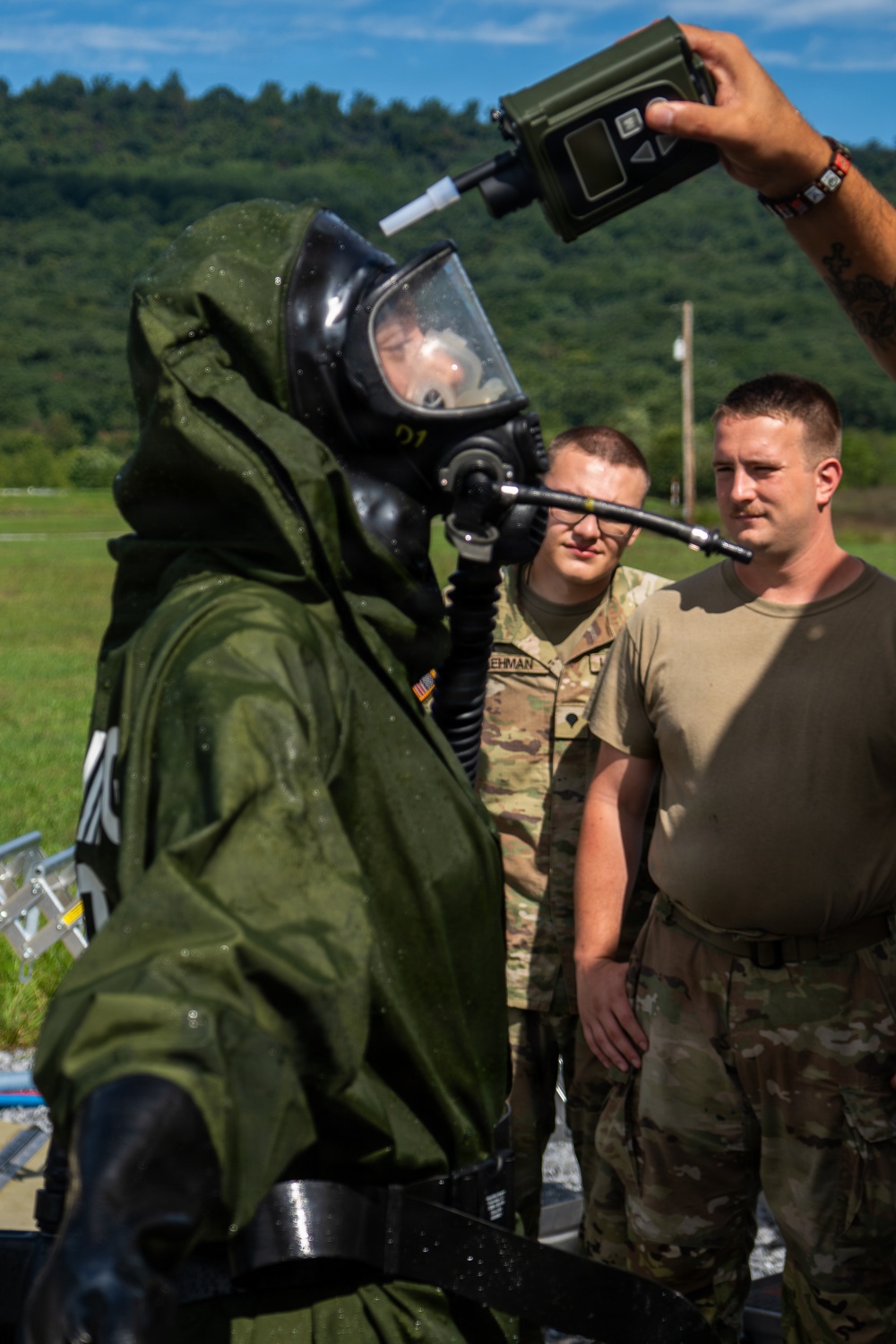 Military personnel are conducting a CBRN (Chemical, Biological, Radiological, and Nuclear) training exercise outdoors. One soldier is wearing a full protective suit with an integrated gas mask and breathing apparatus, while two other soldiers in standard combat uniforms observe. A handheld detection device is being held near the suited soldier's head, likely for decontamination verification or hazard detection.