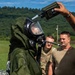 Military personnel are conducting a CBRN (Chemical, Biological, Radiological, and Nuclear) training exercise outdoors. One soldier is wearing a full protective suit with an integrated gas mask and breathing apparatus, while two other soldiers in standard combat uniforms observe. A handheld detection device is being held near the suited soldier's head, likely for decontamination verification or hazard detection.
