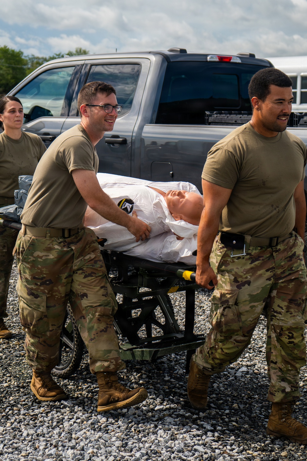 Two military personnel in OCP (Operational Camouflage Pattern) uniforms are transporting a medical training mannequin on a wheeled stretcher across a gravel surface. A third service member is visible in the background. The scene appears to be a medical training exercise, as evidenced by the realistic patient simulator mannequin draped in white.