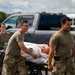 Two military personnel in OCP (Operational Camouflage Pattern) uniforms are transporting a medical training mannequin on a wheeled stretcher across a gravel surface. A third service member is visible in the background. The scene appears to be a medical training exercise, as evidenced by the realistic patient simulator mannequin draped in white.