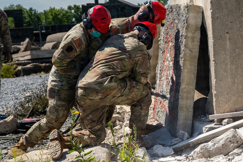 Two U.S. Army soldiers in full combat uniforms and red safety helmets are working together to move or stabilize a large concrete slab at what appears to be a urban search and rescue (USAR) training site. The rubble-strewn environment with broken concrete and debris suggests a simulated collapsed structure exercise. Both personnel are wearing hearing protection and appear to be applying significant physical force to the concrete element.