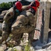 Two U.S. Army soldiers in full combat uniforms and red safety helmets are working together to move or stabilize a large concrete slab at what appears to be a urban search and rescue (USAR) training site. The rubble-strewn environment with broken concrete and debris suggests a simulated collapsed structure exercise. Both personnel are wearing hearing protection and appear to be applying significant physical force to the concrete element.