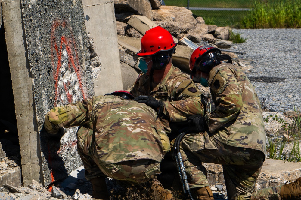 Three personnel in military camouflage uniforms and red safety helmets with face shields are working among concrete rubble, appearing to conduct urban search and rescue or combat engineering operations. They are using pneumatic or hydraulic tools to work on collapsed concrete structures. The scene appears to be a training exercise or disaster response operation at a demolition/rubble site.