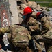 Three personnel in military camouflage uniforms and red safety helmets with face shields are working among concrete rubble, appearing to conduct urban search and rescue or combat engineering operations. They are using pneumatic or hydraulic tools to work on collapsed concrete structures. The scene appears to be a training exercise or disaster response operation at a demolition/rubble site.
