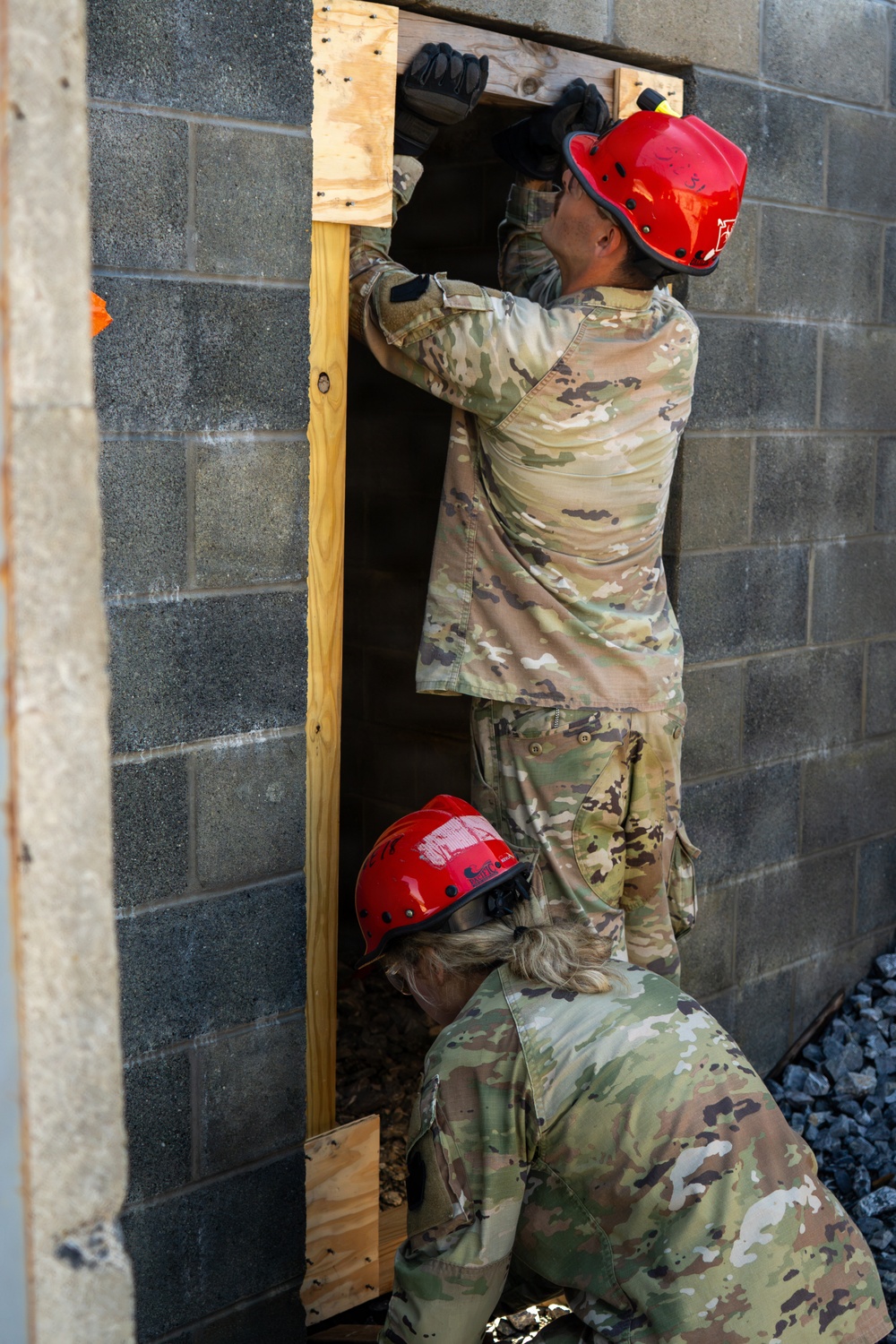 Two military personnel wearing camouflage uniforms and red safety helmets are working together at a narrow opening in a concrete block wall framed with wooden lumber. The standing individual is reaching upward into the framed opening while the second person crouches at the base. This appears to be a search and rescue or urban operations training exercise involving breaching or shoring a structural opening.