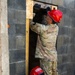 Two military personnel wearing camouflage uniforms and red safety helmets are working together at a narrow opening in a concrete block wall framed with wooden lumber. The standing individual is reaching upward into the framed opening while the second person crouches at the base. This appears to be a search and rescue or urban operations training exercise involving breaching or shoring a structural opening.