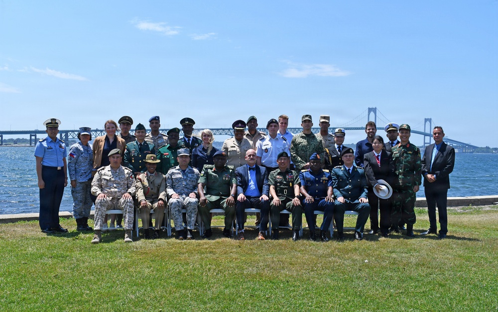 A large group of approximately 25-30 individuals, predominantly in various military uniforms from multiple nations, pose for a formal group photograph outdoors. They are arranged in two rows — seated in the front and standing in the back — on a grassy waterfront area. A large suspension bridge is visible in the background over a body of water, suggesting a coastal or bay location.