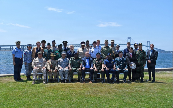 A large group of approximately 25-30 individuals, predominantly in various military uniforms from multiple nations, pose for a formal group photograph outdoors. They are arranged in two rows — seated in the front and standing in the back — on a grassy waterfront area. A large suspension bridge is visible in the background over a body of water, suggesting a coastal or bay location.