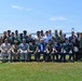 A large group of approximately 25-30 individuals, predominantly in various military uniforms from multiple nations, pose for a formal group photograph outdoors. They are arranged in two rows — seated in the front and standing in the back — on a grassy waterfront area. A large suspension bridge is visible in the background over a body of water, suggesting a coastal or bay location.