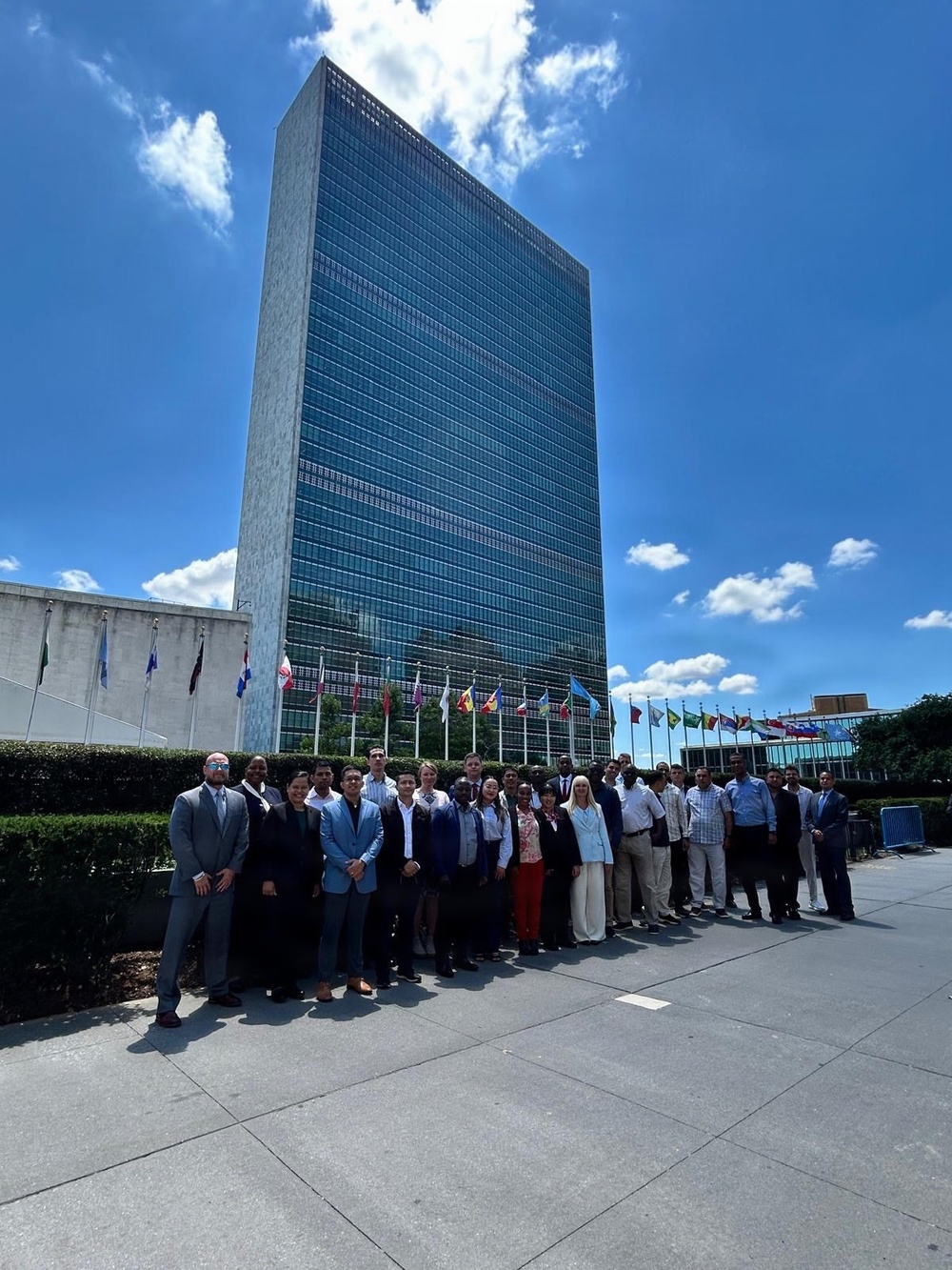 A large group of approximately 25-30 professionally dressed individuals pose for a group photograph in front of the United Nations Secretariat Building in New York City. Multiple national flags are visible in the background along the UN plaza, indicating this is the official UN headquarters complex. The photo appears to be taken on a clear, sunny day with blue skies and scattered clouds.