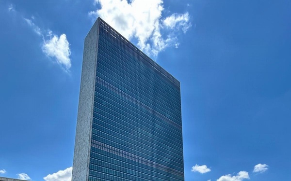 A large group of approximately 25-30 professionally dressed individuals pose for a group photograph in front of the United Nations Secretariat Building in New York City. Multiple national flags are visible in the background along the UN plaza, indicating this is the official UN headquarters complex. The photo appears to be taken on a clear, sunny day with blue skies and scattered clouds.