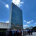 A large group of approximately 25-30 professionally dressed individuals pose for a group photograph in front of the United Nations Secretariat Building in New York City. Multiple national flags are visible in the background along the UN plaza, indicating this is the official UN headquarters complex. The photo appears to be taken on a clear, sunny day with blue skies and scattered clouds.