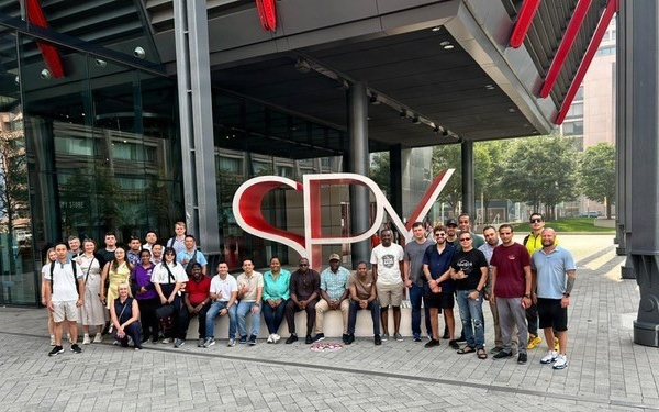 A large group of approximately 30 diverse individuals poses for a group photo in front of a modern building featuring a large red 'SPX' logo sculpture. The setting appears to be an urban commercial or corporate campus with glass-facade architecture, paved plazas, and trees visible in the background. The group appears to be on an organized visit or tour, with members standing and sitting around the large signage.