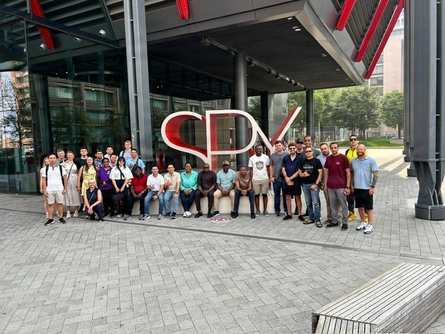 A large group of approximately 30 diverse individuals poses for a group photo in front of a modern building featuring a large red 'SPX' logo sculpture. The setting appears to be an urban commercial or corporate campus with glass-facade architecture, paved plazas, and trees visible in the background. The group appears to be on an organized visit or tour, with members standing and sitting around the large signage.