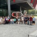 A large group of approximately 30 diverse individuals poses for a group photo in front of a modern building featuring a large red 'SPX' logo sculpture. The setting appears to be an urban commercial or corporate campus with glass-facade architecture, paved plazas, and trees visible in the background. The group appears to be on an organized visit or tour, with members standing and sitting around the large signage.