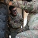 Two U.S. Army soldiers in Operational Camouflage Pattern (OCP) uniforms are performing maintenance on a large military vehicle tire. One soldier, whose name tape reads partially as 'TON' and wears a 1st Cavalry Division patch, is wearing safety goggles and working on the wheel hub area. They appear to be conducting vehicle maintenance or a tire change in what looks like a motor pool or maintenance bay.