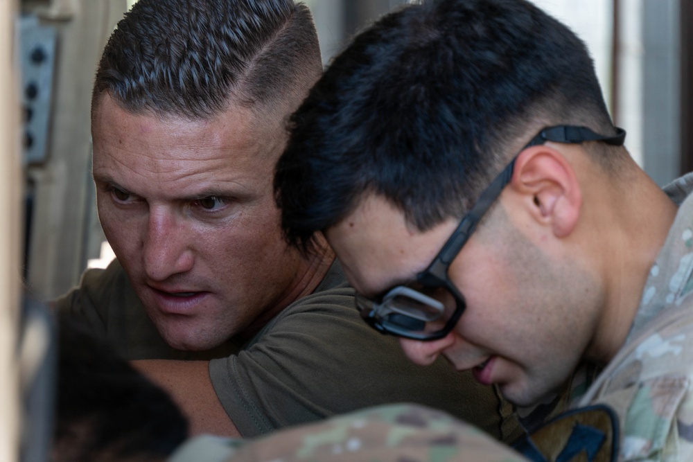 Two men are closely examining or working on something together, with focused expressions. One man wears a olive/green t-shirt and the other wears a military multicam/camouflage uniform with tactical eyewear pushed up on his head. The close-up framing and intensity of their focus suggests a technical task or briefing is taking place.