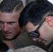 Two men are closely examining or working on something together, with focused expressions. One man wears a olive/green t-shirt and the other wears a military multicam/camouflage uniform with tactical eyewear pushed up on his head. The close-up framing and intensity of their focus suggests a technical task or briefing is taking place.