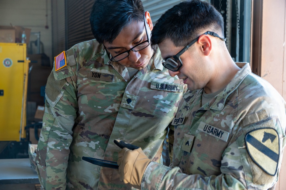 Two U.S. Army soldiers in OCP (Operational Camouflage Pattern) uniforms are reviewing content on a handheld device, likely a tablet or smartphone. One soldier, identified by name tape as 'YON,' wears black-framed glasses, while the other displays a 1st Cavalry Division patch on his right shoulder sleeve. They appear to be in a warehouse or depot environment.