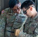 Two U.S. Army soldiers in OCP (Operational Camouflage Pattern) uniforms are reviewing content on a handheld device, likely a tablet or smartphone. One soldier, identified by name tape as 'YON,' wears black-framed glasses, while the other displays a 1st Cavalry Division patch on his right shoulder sleeve. They appear to be in a warehouse or depot environment.