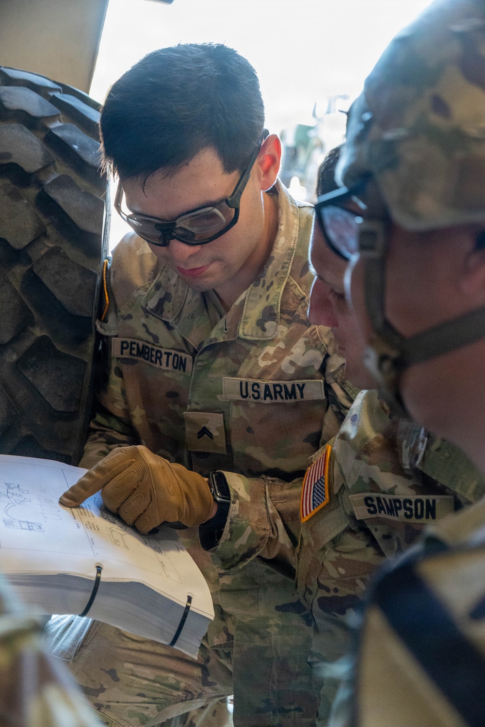 Two U.S. Army soldiers in OCP (Operational Camouflage Pattern) uniforms are reviewing a document or technical manual together. The soldier in the foreground, identified by name tape as 'PEMBERTON,' is pointing at the document while wearing tactical gloves and protective eyewear. A second soldier identified as 'SAMPSON' is visible on the right side of the frame.