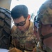 Two U.S. Army soldiers in OCP (Operational Camouflage Pattern) uniforms are reviewing a document or technical manual together. The soldier in the foreground, identified by name tape as 'PEMBERTON,' is pointing at the document while wearing tactical gloves and protective eyewear. A second soldier identified as 'SAMPSON' is visible on the right side of the frame.