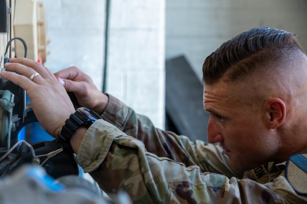 A male soldier in military camouflage uniform is closely inspecting or working on a piece of equipment or panel. He appears focused and concentrated on a technical task, leaning in closely to examine components. The image is a close-up shot emphasizing his face and hands.