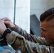 A male soldier in military camouflage uniform is closely inspecting or working on a piece of equipment or panel. He appears focused and concentrated on a technical task, leaning in closely to examine components. The image is a close-up shot emphasizing his face and hands.