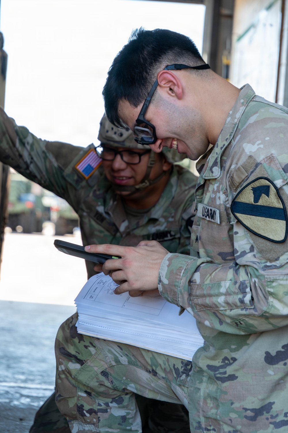 Two U.S. Army soldiers in MultiCam/OCP camouflage uniforms are reviewing paperwork together, with one soldier holding a mobile device while referencing a stack of documents. The foreground soldier wears a 1st Cavalry Division shoulder sleeve insignia and a 'U.S. ARMY' tape. They appear to be conducting administrative or logistical work in what looks like a loading dock or vehicle bay area.