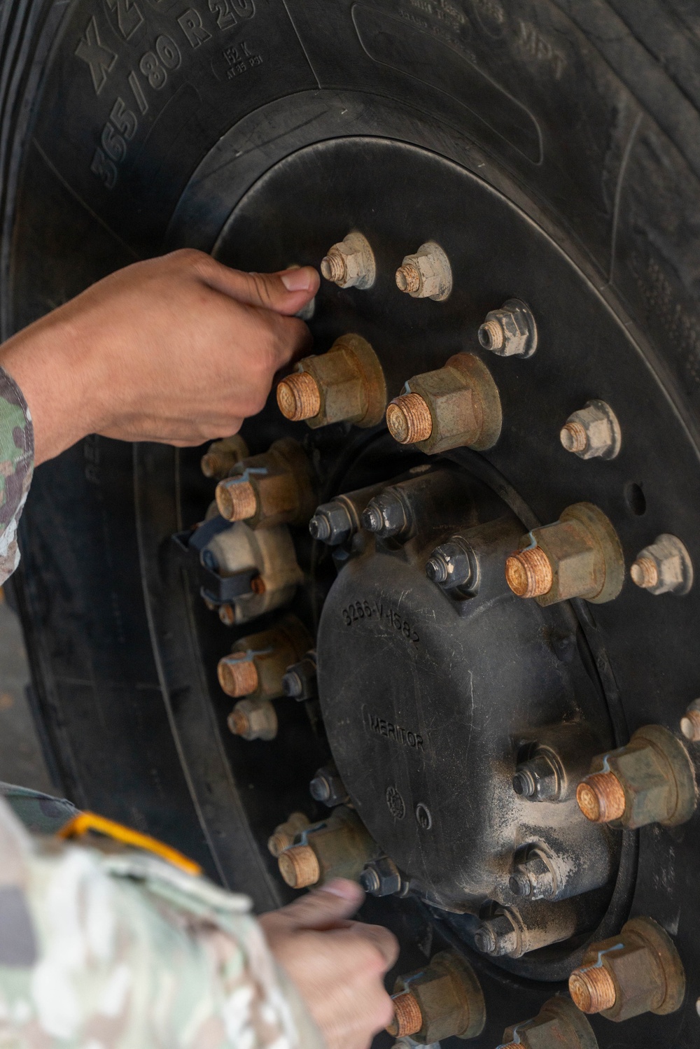 A person in military camouflage uniform is inspecting or working on a large vehicle wheel assembly. The image shows a close-up of a heavy-duty tire and hub with multiple lug nuts and bolts, featuring a Meritor brand hub component. The wheel appears to belong to a large military or heavy transport vehicle.