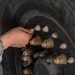 A person in military camouflage uniform is inspecting or working on a large vehicle wheel assembly. The image shows a close-up of a heavy-duty tire and hub with multiple lug nuts and bolts, featuring a Meritor brand hub component. The wheel appears to belong to a large military or heavy transport vehicle.