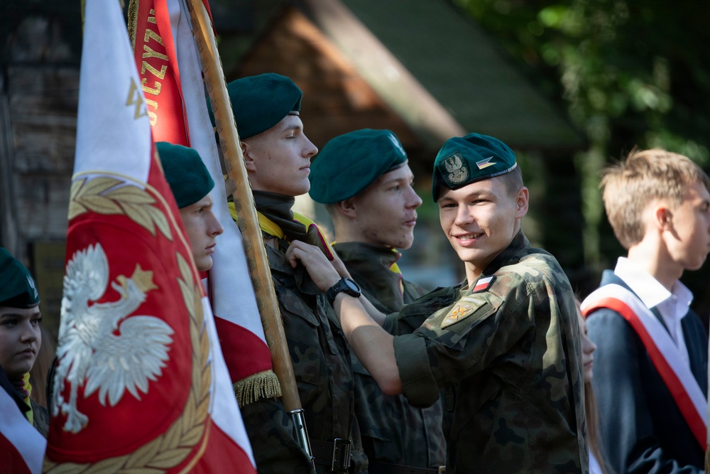 A group of young people in military-style camouflage uniforms and dark green berets are participating in a formal ceremony, likely in Poland based on the visible Polish national flag and eagle emblem. One smiling youth appears to be adjusting or pinning something on another's uniform. The setting appears to be an outdoor ceremonial event with a wooden structure visible in the background.