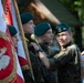 A group of young people in military-style camouflage uniforms and dark green berets are participating in a formal ceremony, likely in Poland based on the visible Polish national flag and eagle emblem. One smiling youth appears to be adjusting or pinning something on another's uniform. The setting appears to be an outdoor ceremonial event with a wooden structure visible in the background.