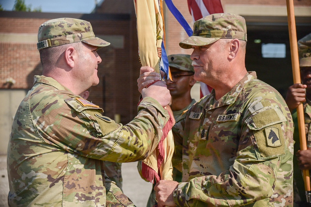 Two U.S. Army soldiers in OCP (Operational Camouflage Pattern) uniforms are participating in a formal guidon transfer ceremony, likely a change of command or responsibility. The soldier on the right, identified by the name tape 'PARTIN' and wearing a First Cavalry Division patch, is passing or receiving a unit guidon to/from the soldier on the left. Additional soldiers in uniform are visible in the background holding flags.